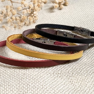 Può includere: Quattro braccialetti in pelle nei colori rosso, giallo, marrone e nero, ciascuno con una chiusura argentata. I braccialetti sono impilati su una superficie strutturata bianco sporco, con fiori secchi sullo sfondo.