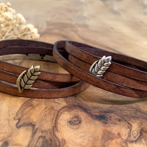 Può includere: Due braccialetti avvolgenti in pelle marrone con chiusure in metallo a forma di spiga di grano. I braccialetti sono a strati e sembrano essere realizzati in vera pelle. Le chiusure a forma di grano sono in tonalità oro e argento. I braccialetti sono esposti su una superficie di legno.