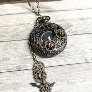 May include: A steampunk-style pocket watch pendant necklace. The watch features a black face with visible gears and a key charm. The pendant is attached to a silver chain, with additional gear and arrow charms.