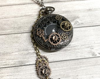 May include: A steampunk-style pocket watch pendant necklace. The watch features a black face with visible gears and a key charm. The pendant is attached to a silver chain, with additional gear and arrow charms.
