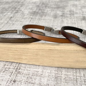 Può includere: Tre braccialetti in pelle con chiusura argentata. I braccialetti sono marroni, marrone scuro e grigi.
