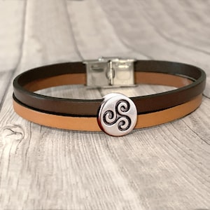 Pode incluir: Pulseira de couro de duas camadas com fecho prateado e berloque decorativo prateado. A pulseira apresenta uma camada superior castanho escuro e uma camada inferior castanho claro. O berloque tem um design de espiral celta.
