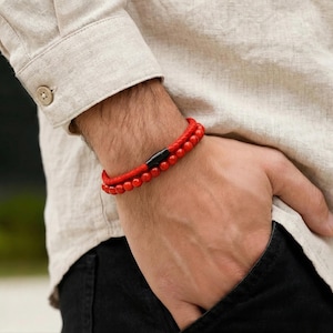Peut inclure: Un bracelet rouge à perles avec un fermoir magnétique noir est porté au poignet. Le bracelet est composé de petites perles rondes. La personne porte une chemise boutonnée beige clair et un pantalon noir.