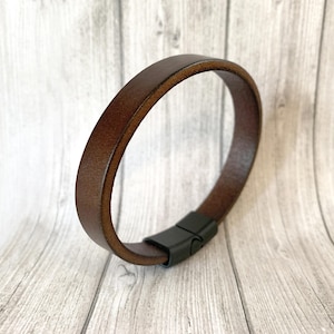 May include: A brown leather bracelet with a black magnetic clasp. The bracelet is a simple band design, with a smooth, polished finish. The leather appears to be a rich, dark brown color.