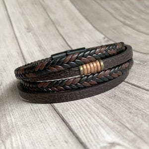 Könnte beinhalten: Ein geflochtenes Lederarmband mit braunem und schwarzem Design. Das Armband hat einen Metallverschluss und eine dekorative Metallperle.