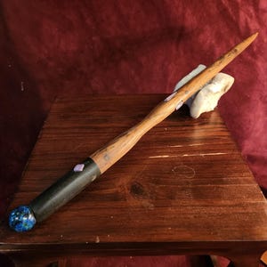 May include: A handcrafted wooden wand with a dark handle and a blue and white orb at the base. The wand's shaft is a light brown color with purple accents. The wand is resting on a wooden table.