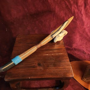 May include: A handcrafted wooden spear with a pointed tip, featuring a light-colored section and a wrapped copper wire section. The spear is resting on a small, dark wooden table, with a piece of light-colored stone.