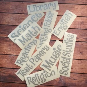 May include: A set of ten gray vinyl labels with various school subjects written on them, including "Library", "Science", "Math", "Reading", "Test Papers", "Writing Folder", "English", "Music", "Religion", and "Social Studies".