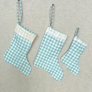 May include: Three Christmas stockings in varying sizes, each featuring a light blue and white gingham check pattern. Every stocking is accented with a white lace trim along the top cuff and includes a matching fabric loop for hanging purposes.