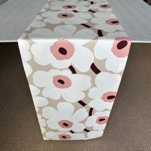 May include: A rectangular table runner featuring a repeating floral pattern with large white petals, soft pink centres, and dark maroon circular cores set against a light beige background. The fabric displays a smooth texture and clean edges.