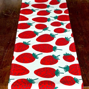 May include: A rectangular table runner featuring a repeating pattern of bright red strawberries with green leafy stems on a solid white background. The strawberries are oriented in various directions across the fabric surface, creating a consistent graphic print.