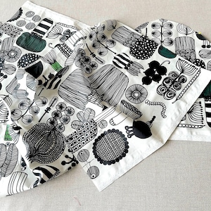 Peut inclure: Tissu blanc avec un motif botanique noir et vert. Le motif présente des formes végétales stylisées, notamment des fleurs, des feuilles et des formes géométriques. Le tissu est plié, montrant la répétition du motif.