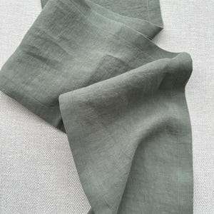 May include: A sage green linen table runner with a subtle texture. The fabric is folded and draped, showcasing the natural weave and soft color. The edges are neatly hemmed, adding a tailored finish. Ideal for adding a touch of elegance to any dining table.