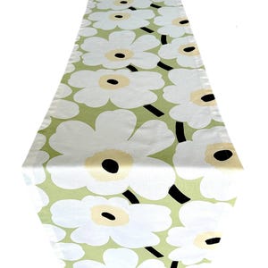 May include: A white table runner with a repeating floral pattern. The flowers have white petals, light yellow centers, and black accents. The background is a light green color.