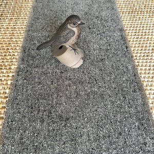 May include: A small, brown and grey ceramic bird figurine perched on a light tan, cylindrical base. The bird features detailed feather textures and a dark beak. It is positioned on a rectangular, grey, textured fabric runner with a woven pattern.