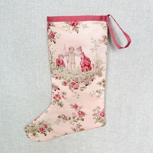 May include: A pale pink Christmas stocking featuring a toile de jouy pattern with red floral motifs and a scene of two children and a dog. The stocking has a solid pink ribbon trim at the top and a matching fabric loop for hanging on a hook.