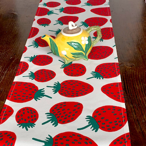 May include: A white table runner with a repeating pattern of large red strawberries with green stems. A yellow teapot with green handle and floral accents sits on the runner.