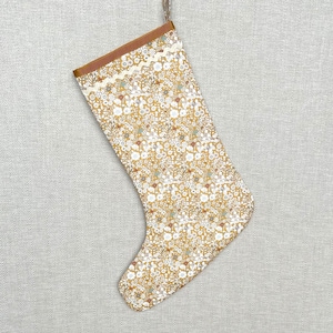 May include: A Christmas stocking featuring a mustard yellow background with a dense floral pattern of white, brown, and muted green flowers. The top cuff is a solid brown fabric accented with a white rickrack trim and a small hanging loop.