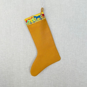 May include: A mustard yellow Christmas stocking featuring a decorative cuff with a colourful pattern of blue horses, red animals, and green floral shapes. The stocking includes a silver hanging loop at the top for display purposes during the holidays.