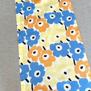 May include: A table runner with a floral pattern. The design features large, stylized flowers in blue, orange, and yellow against a white backdrop. The flowers have dark brown centres and stems. The runner has a yellow border.