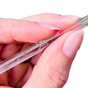 May include: A clear glass nail pusher with a silicone tip. The pusher is held in a hand.
