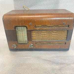Peut inclure: Poste de radio vintage en bois avec une finition marron foncé. La radio a une forme rectangulaire aux bords arrondis et une grille de haut-parleur recouverte d'un tissu beige. Elle comporte un cadran de réglage et deux boutons. Un fin fil jaune dépasse du haut.