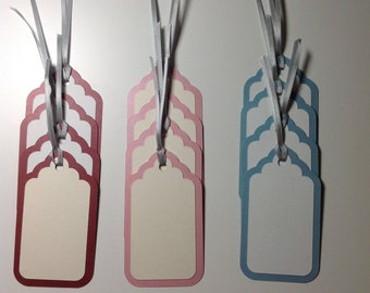 May include: Set of 15 gift tags with scalloped edges. The tags are in three colors: burgundy, pink, and light blue. Each tag has a white center and a silver ribbon.