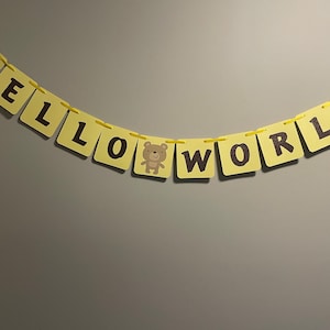 Peut inclure: Une bannière jaune avec les mots "HELLO WORLD" en lettres noires. Un petit ours en peluche brun se trouve entre le "O" et le "W".