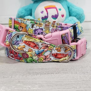 May include: Two pink dog collars with a colorful Care Bears print. The collars feature a pink buckle and a silver D-ring. The print includes Care Bears, crayons, and other colorful objects.