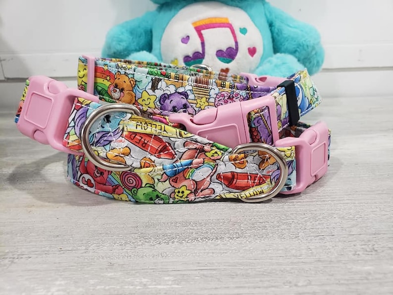 May include: Two pink dog collars with a colorful Care Bears print. The collars feature a pink buckle and a silver D-ring. The print includes Care Bears, crayons, and other colorful objects.