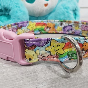 May include: A pink and white dog collar with a Care Bears print featuring rainbows, stars, and Care Bears characters. The collar has a pink plastic buckle and a silver metal D-ring.