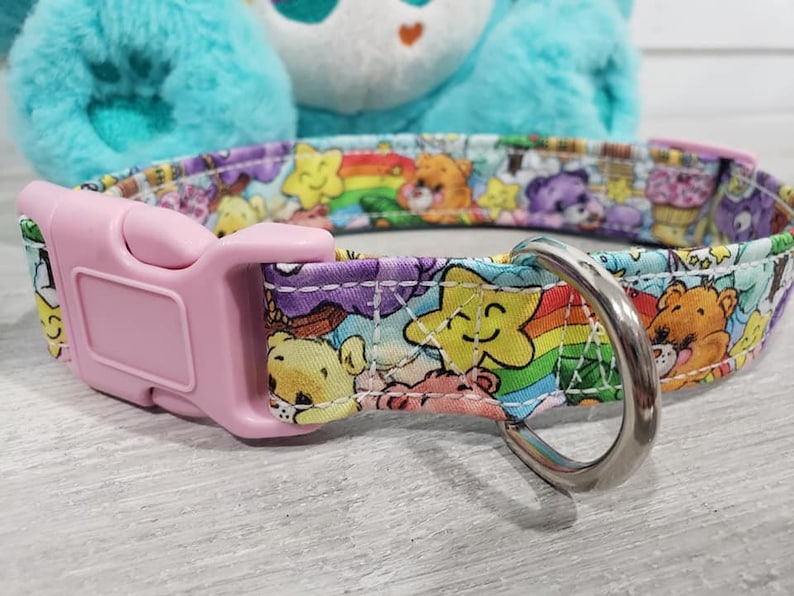 May include: A pink and white dog collar with a Care Bears print featuring rainbows, stars, and Care Bears characters. The collar has a pink plastic buckle and a silver metal D-ring.