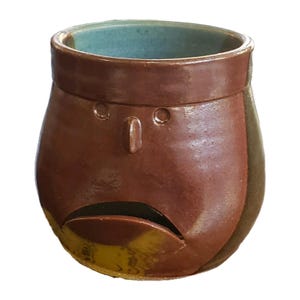 May include: A handmade ceramic mug with a face design. The mug is primarily brown with a teal interior and yellow and green accents. The face features a nose, eyes, and a wide open mouth. This unique mug is perfect for coffee or tea.
