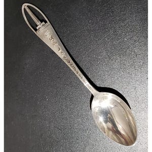 May include: A silver-toned metal souvenir spoon featuring a decorative handle with an open-work top and the embossed text "LAS VEGAS" running down the stem. The spoon has a smooth, reflective oval bowl and a tapered handle design.