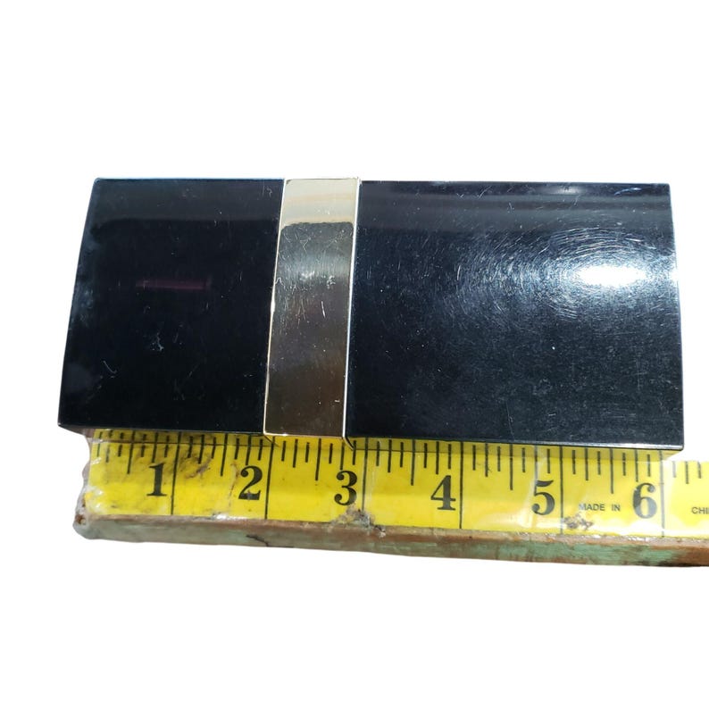Puede incluir: Un tarjetero rectangular negro con un acento de metal plateado. El soporte mide aproximadamente 14 cm de largo, medido con una cinta m&eacute;trica amarilla.
