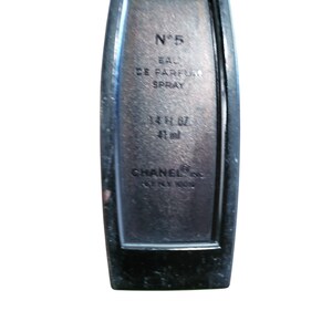 Puede incluir: Un frasco de perfume negro con el texto "N&deg;5 EAU DE PARFUM SPRAY 42 ml CHANEL PARIS NEW YORK". El frasco tiene forma rectangular con bordes redondeados y un acabado oscuro y met&aacute;lico.