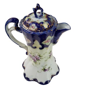 May include: An antique porcelain coffee pot with a navy blue and white floral design. The pot features a curved handle, a spout, and a lid with a decorative finial. Gold accents highlight the intricate details of the flowers and patterns.