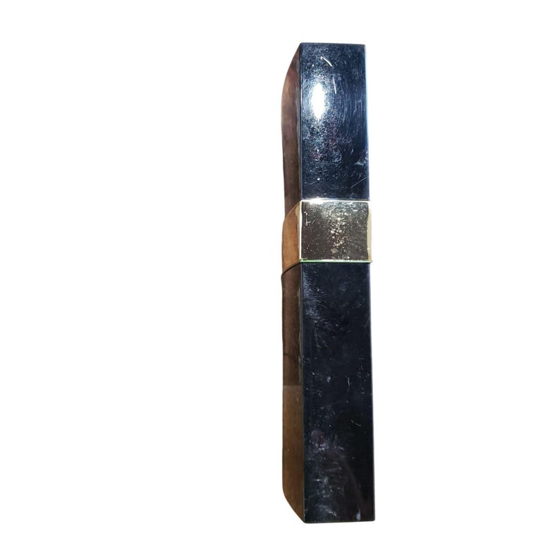 Puede incluir: Un tubo de l&aacute;piz labial negro con una banda dorada. El envase del l&aacute;piz labial es rectangular y tiene una superficie brillante y reflectante. La banda dorada se encuentra cerca del centro del tubo.