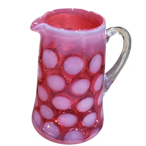 May include: A pink glass pitcher with a clear handle and spout. The pitcher has a pattern of large, light pink circles against a darker pink background. The pitcher is likely used for serving beverages.