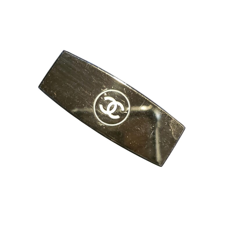 Puede incluir: Pinza para el pelo rectangular plateada con un logotipo blanco circular en el centro. El logotipo presenta dos C entrelazadas en espejo. La pinza tiene una forma ligeramente curvada y un acabado brillante.