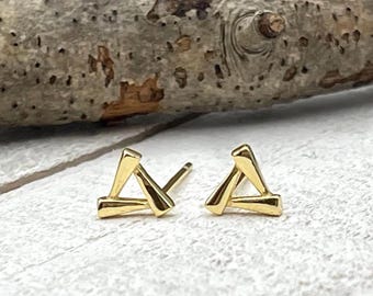 May include: A pair of gold-colored triangle stud earrings. The earrings are small and geometric, with a simple, modern design. They are displayed on a white surface with a wooden branch in the background.