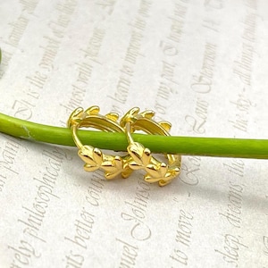 May include: A pair of gold-colored hoop earrings. Each earring features a leaf design around the hoop. The earrings are resting on a green stem against a background of printed text.