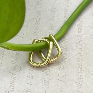 May include: A pair of gold-colored, geometric hoop earrings. The earrings are a unique, angular shape, and are displayed on a green stem against a backdrop of printed text.