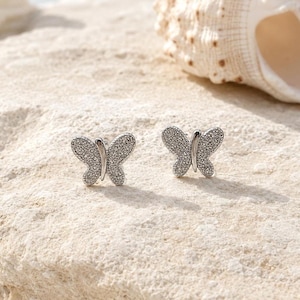 May include: A pair of silver-toned stud earrings shaped like butterflies. Each earring features a smooth central body and wings covered in small, clear, sparkling stones that catch the light, creating a textured and reflective surface appearance.
