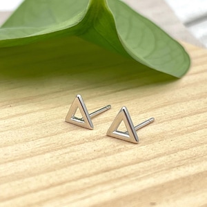 May include: A pair of silver triangle stud earrings. The earrings have a minimalist design. They are displayed on a light brown wooden surface, with a green leaf in the background. The earrings are small and geometric.