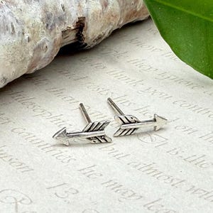 May include: A pair of silver arrow stud earrings. The earrings are detailed with feather-like designs and have a post backing. The earrings are displayed on a textured, off-white surface with faint text.