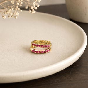 Peut inclure: Une paire de petites créoles dorées ornées d'une rangée de pierres circulaires rose vif incrustées sur la face avant. Les boucles d'oreilles sont de forme circulaire avec un mécanisme de fermeture à charnière, présentant une finition métallique polie.