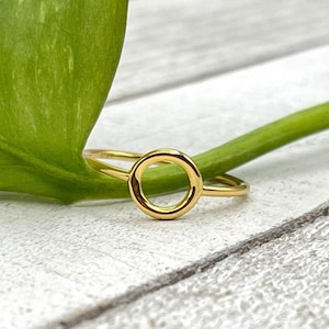May include: A gold-colored ring with a circular design. The ring is set against a background of a green leaf and a white wooden surface. The ring's circular element is the focal point.