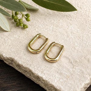 May include: A pair of gold-toned hoop earrings featuring an elongated, rectangular oval shape with rounded corners. The earrings have a polished, reflective metallic finish and a hinged closure mechanism, presenting a clean and simple design.