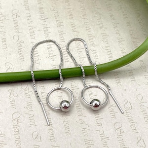 May include: A pair of silver threader earrings. Each earring features a small, polished silver ball suspended within a silver hoop. A long, delicate chain extends from the hoop, creating a minimalist and elegant design.